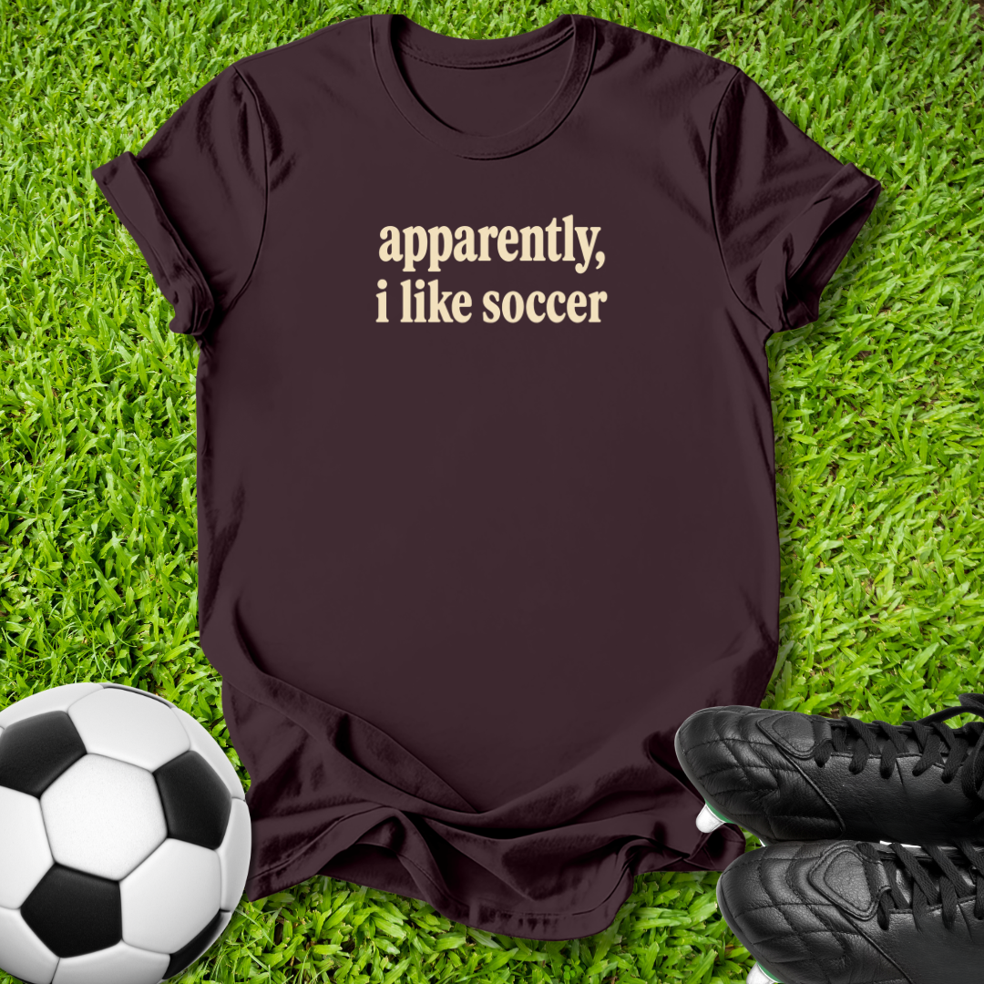 A dark brown short-sleeve t-shirt with the text 'apparently, i like soccer' printed in beige on the chest, laid flat on green grass with a soccer ball and black cleats as props.