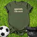 An olive green short-sleeve t-shirt with the text 'apparently, i like soccer' printed in beige on the chest, laid flat on green grass with a soccer ball and black cleats nearby.