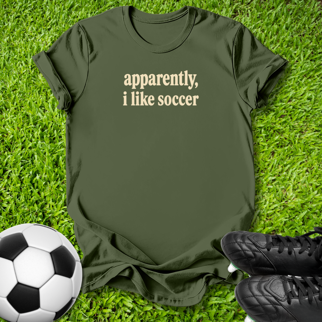 An olive green short-sleeve t-shirt with the text 'apparently, i like soccer' printed in beige on the chest, laid flat on green grass with a soccer ball and black cleats nearby.