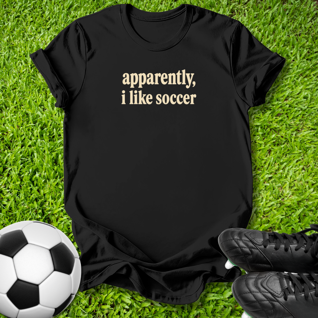 A black short-sleeve t-shirt with the phrase 'apparently, i like soccer' printed in beige serif text, laid flat on green grass with a soccer ball and black cleats nearby.