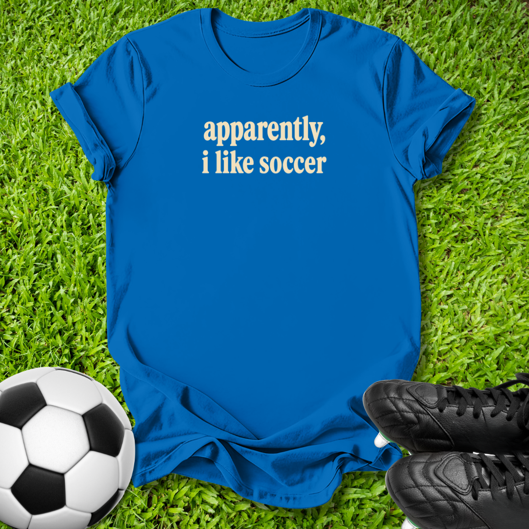 A royal blue short-sleeve t-shirt with the phrase 'apparently, i like soccer' printed in white text across the chest, laid flat on green grass with a soccer ball and black cleats nearby.