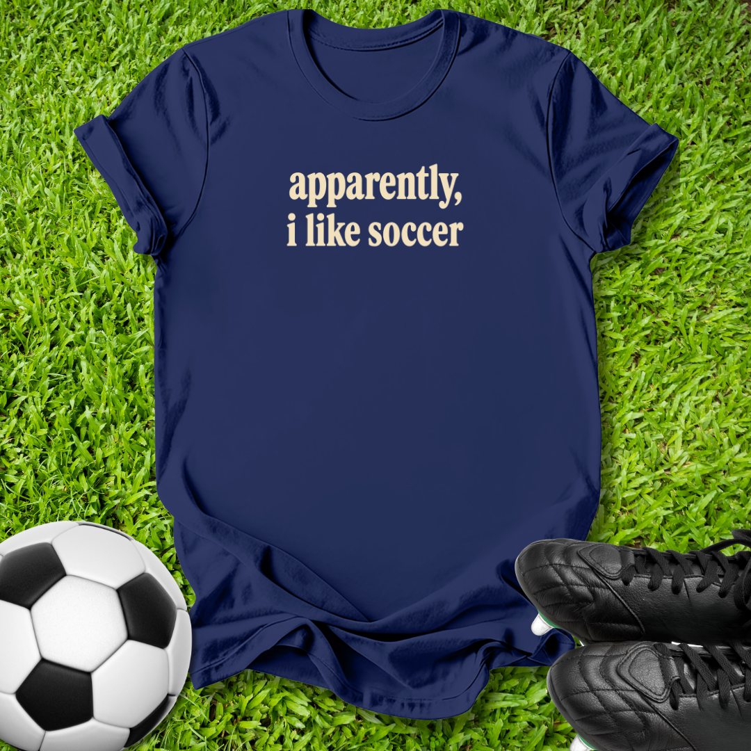 A navy blue short-sleeve t-shirt with the text 'apparently, i like soccer' printed in white serif font, laid flat on green grass with a soccer ball and black cleats nearby.