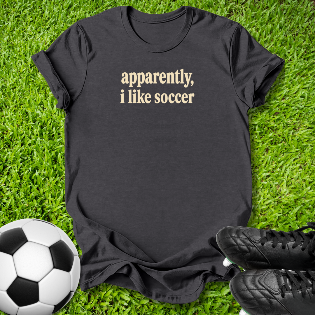 A black short-sleeve t-shirt with the text 'apparently, i like soccer' printed in beige on the chest, laid flat on green grass with a soccer ball and black cleats nearby.