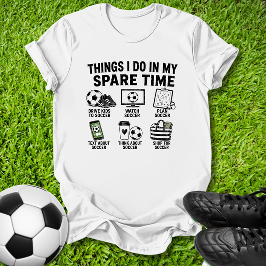 A white graphic t-shirt laid flat on green grass, featuring a black printed design with soccer-themed illustrations and the text 'THINGS I DO IN MY SPARE TIME.' A soccer ball is at the bottom left, and black soccer cleats are at the bottom right.