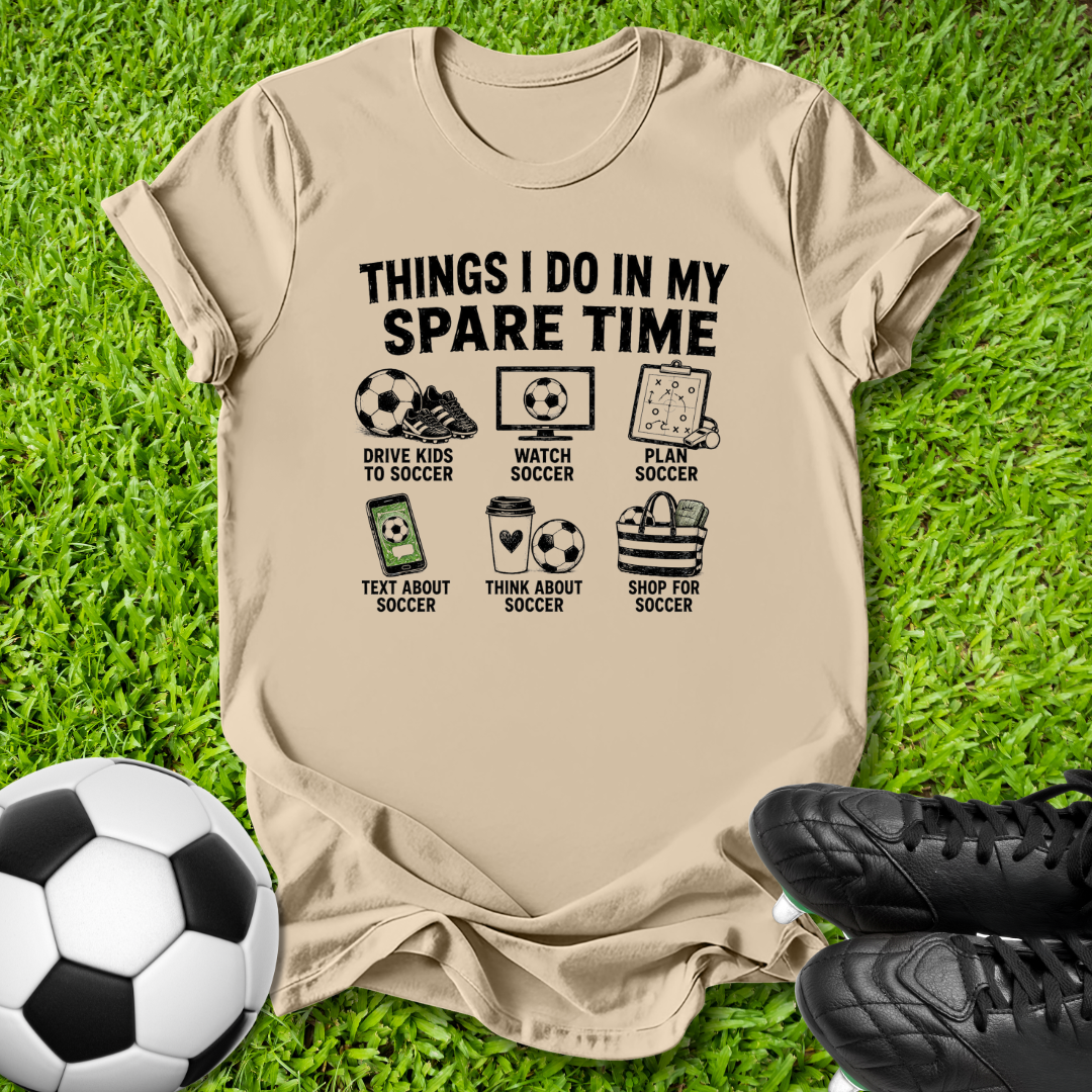 A beige graphic t-shirt laid flat on green grass, featuring black text that reads 'THINGS I DO IN MY SPARE TIME' above soccer-themed illustrations. A soccer ball is at the bottom left and black soccer cleats are at the bottom right.