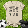 A beige graphic t-shirt laid flat on green grass, featuring a black printed design with soccer-themed icons and the text 'THINGS I DO IN MY SPARE TIME'. A soccer ball is on the left, and black soccer cleats are on the right.