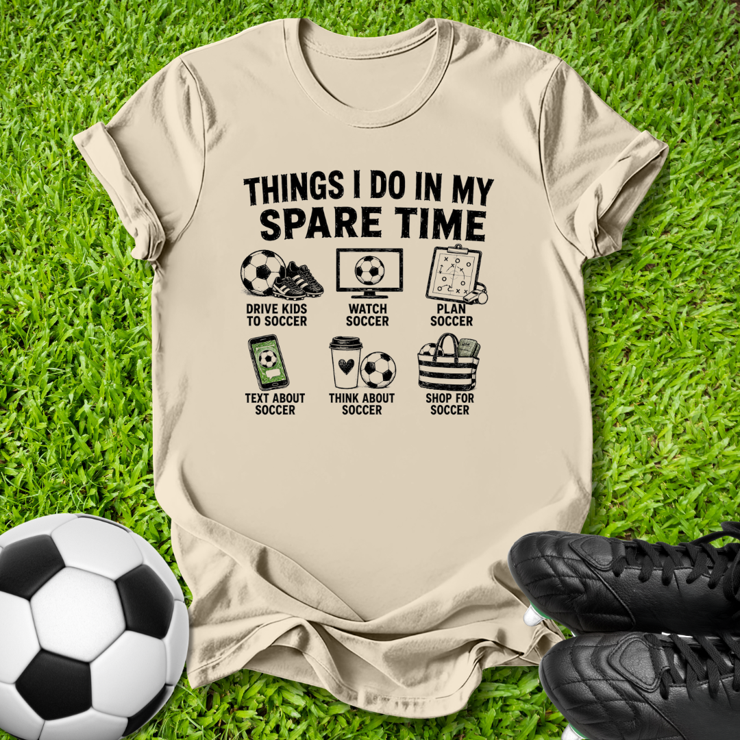 A beige graphic t-shirt laid flat on green grass, featuring a black printed design with soccer-themed icons and the text 'THINGS I DO IN MY SPARE TIME'. A soccer ball is on the left, and black soccer cleats are on the right.