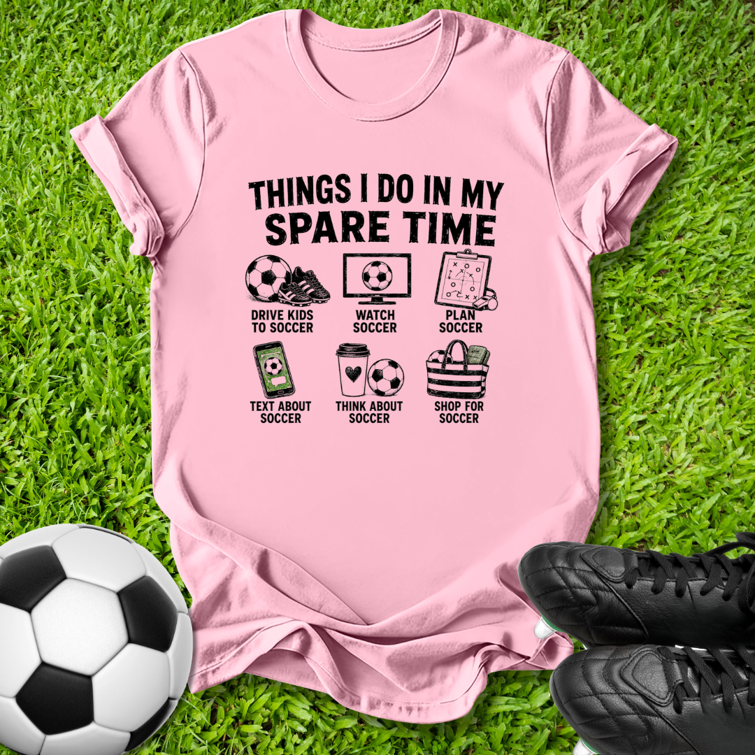 A pink short-sleeve graphic T-shirt laid flat on green grass, with soccer-themed illustrations and text reading 'THINGS I DO IN MY SPARE TIME.' A soccer ball is at the lower left and black soccer cleats are at the lower right.