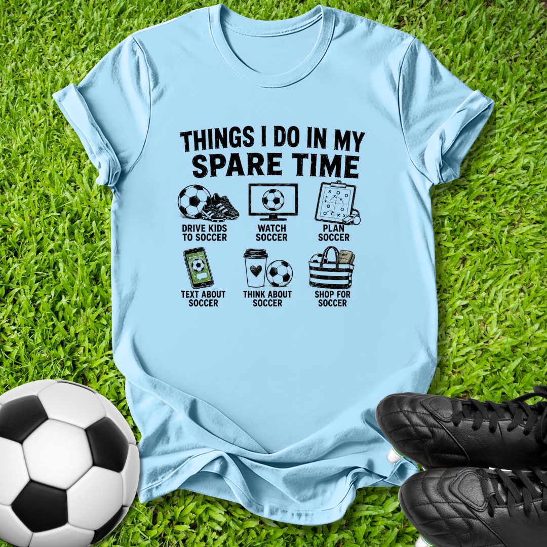 A light blue graphic t-shirt laid flat on green grass, featuring a black printed design with soccer-themed illustrations and the text 'THINGS I DO IN MY SPARE TIME'. A soccer ball is on the left, and black soccer cleats are on the right.
