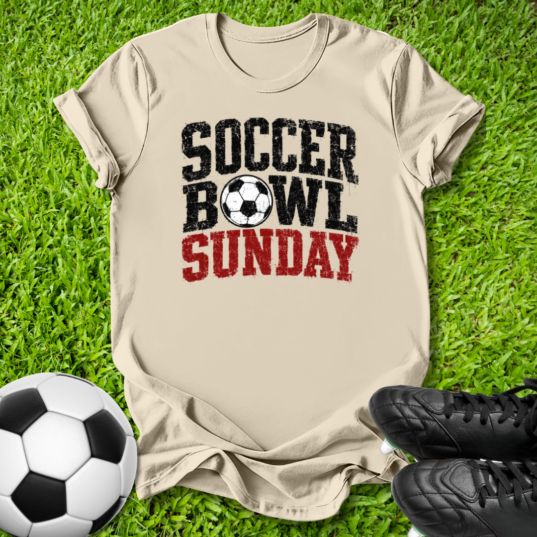 A beige graphic t-shirt laid flat on green grass, featuring the text 'SOCCER BOWL SUNDAY' in black and red with a soccer ball graphic. A soccer ball and black cleats are placed around it as props.