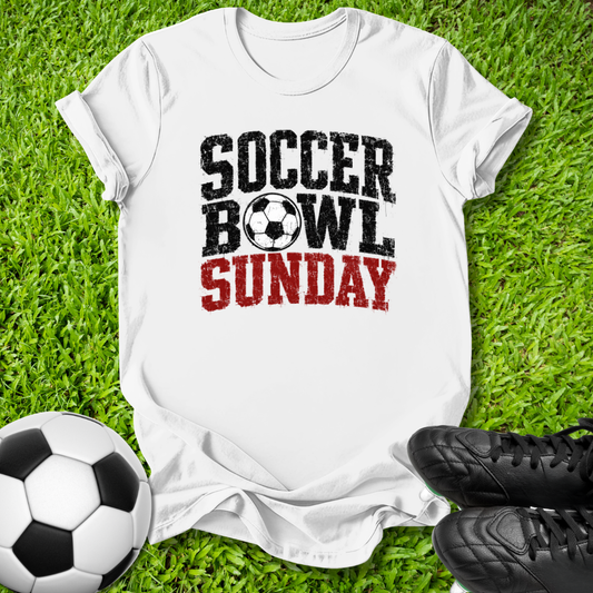 A white short-sleeve graphic T-shirt laid flat on green grass, with a soccer ball at the lower left and black cleats at the lower right. The shirt reads 'SOCCER BOWL SUNDAY' in black and red distressed lettering, with a soccer ball icon between the words.