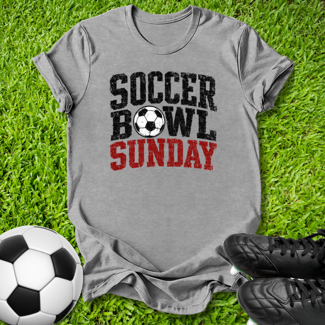 A heather gray graphic t-shirt laid flat on green grass, featuring the text 'SOCCER BOWL SUNDAY' in distressed black and red lettering with a soccer ball graphic. A soccer ball and black cleats are styled around the shirt.