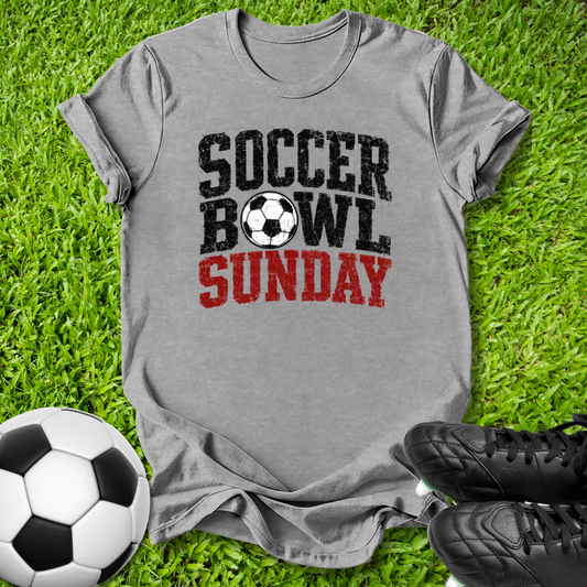 A heather gray graphic t-shirt laid flat on green grass, featuring the text 'SOCCER BOWL SUNDAY' in distressed black and red lettering with a soccer ball graphic. A soccer ball and black cleats are styled around the shirt.