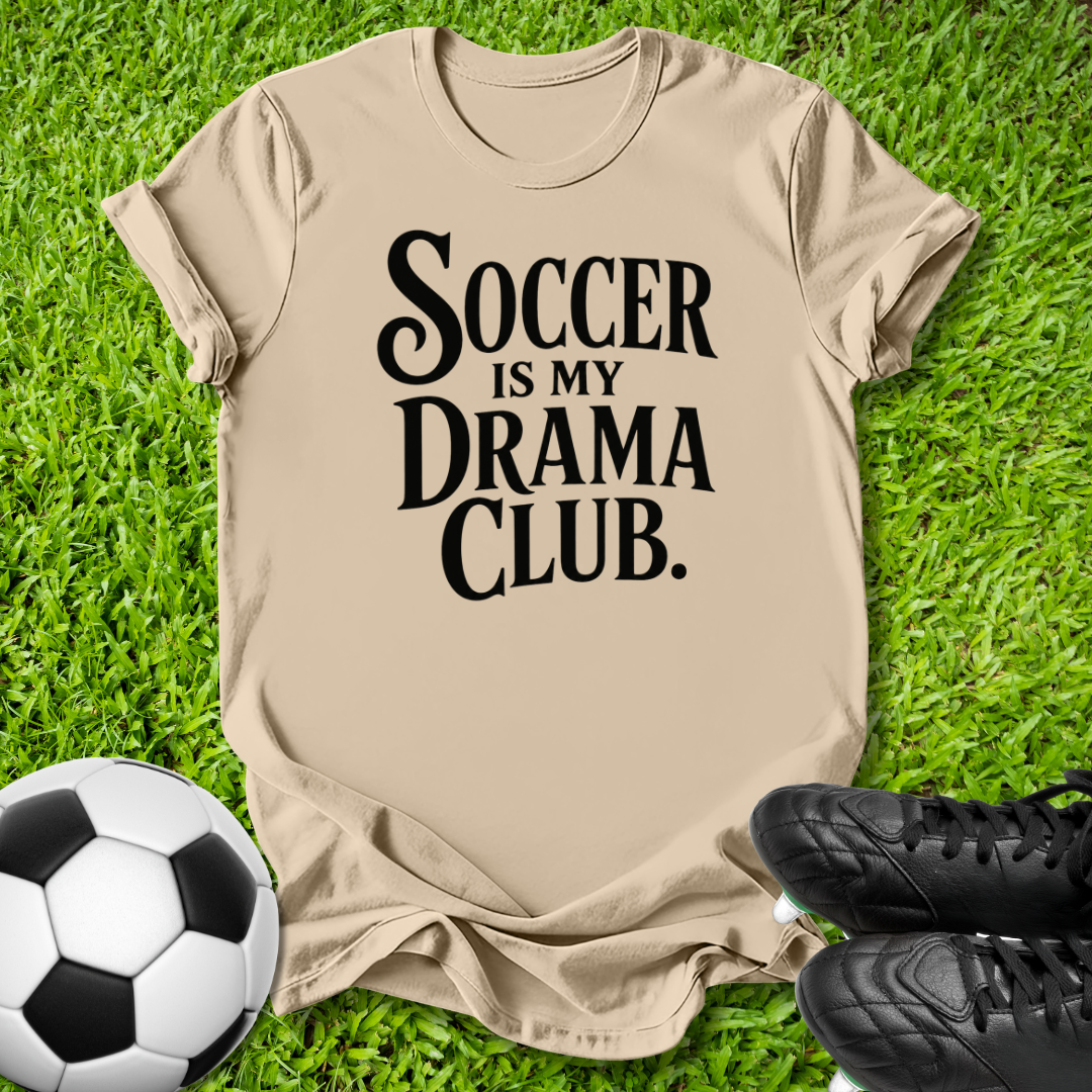 A beige graphic t-shirt laid flat on green grass, featuring the text 'SOCCER IS MY DRAMA CLUB.' in black.