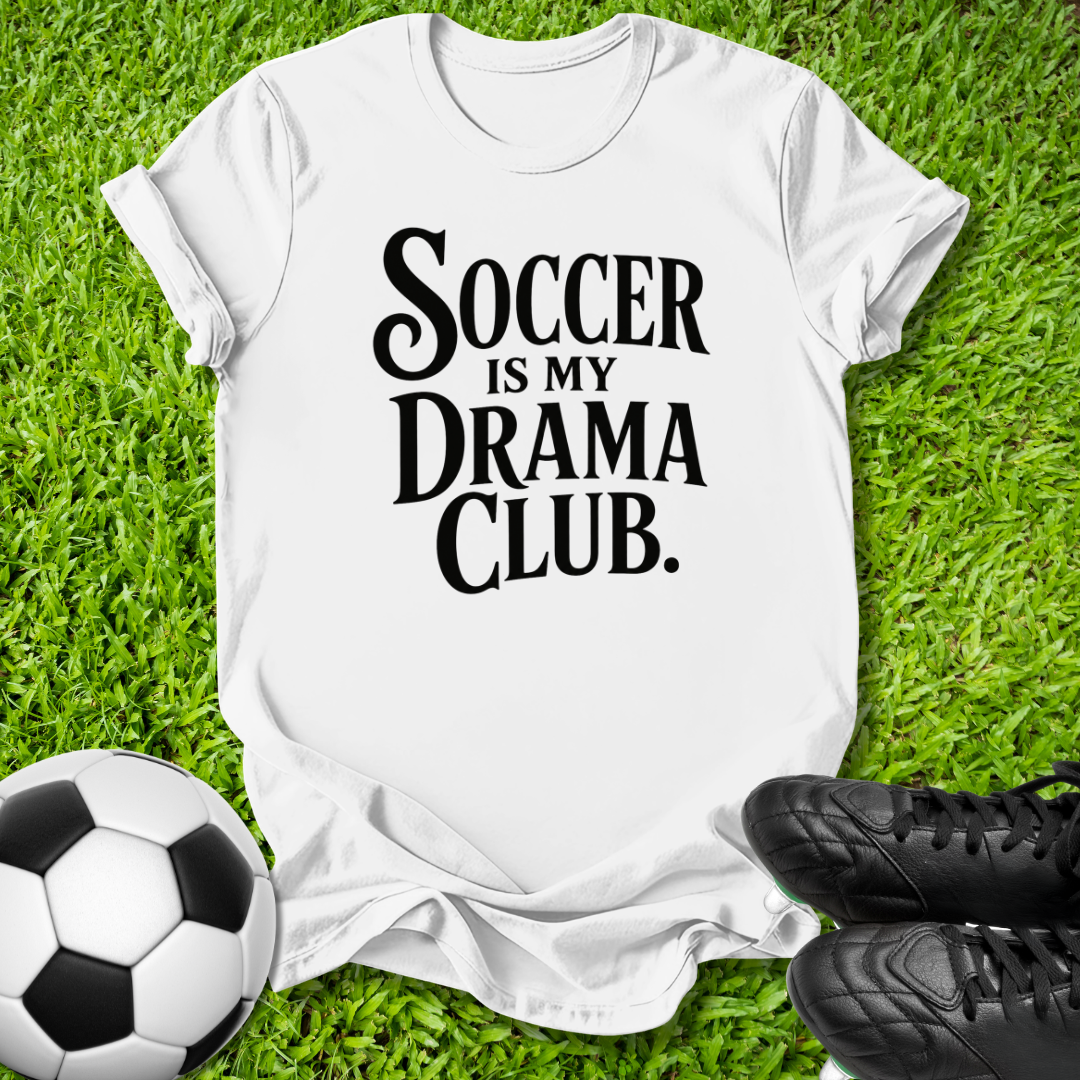 A white short-sleeve T-shirt laid flat on green grass, printed with the phrase 'SOCCER IS MY DRAMA CLUB.' in large black lettering.