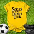A bright yellow graphic t-shirt laid flat on green grass, featuring the text 'SOCCER IS MY DRAMA CLUB.' in black.