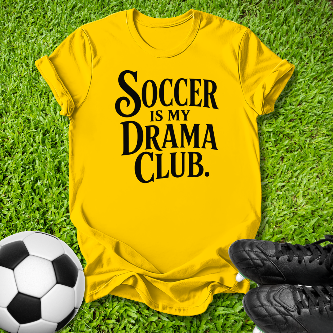 A bright yellow graphic t-shirt laid flat on green grass, featuring the text 'SOCCER IS MY DRAMA CLUB.' in black.