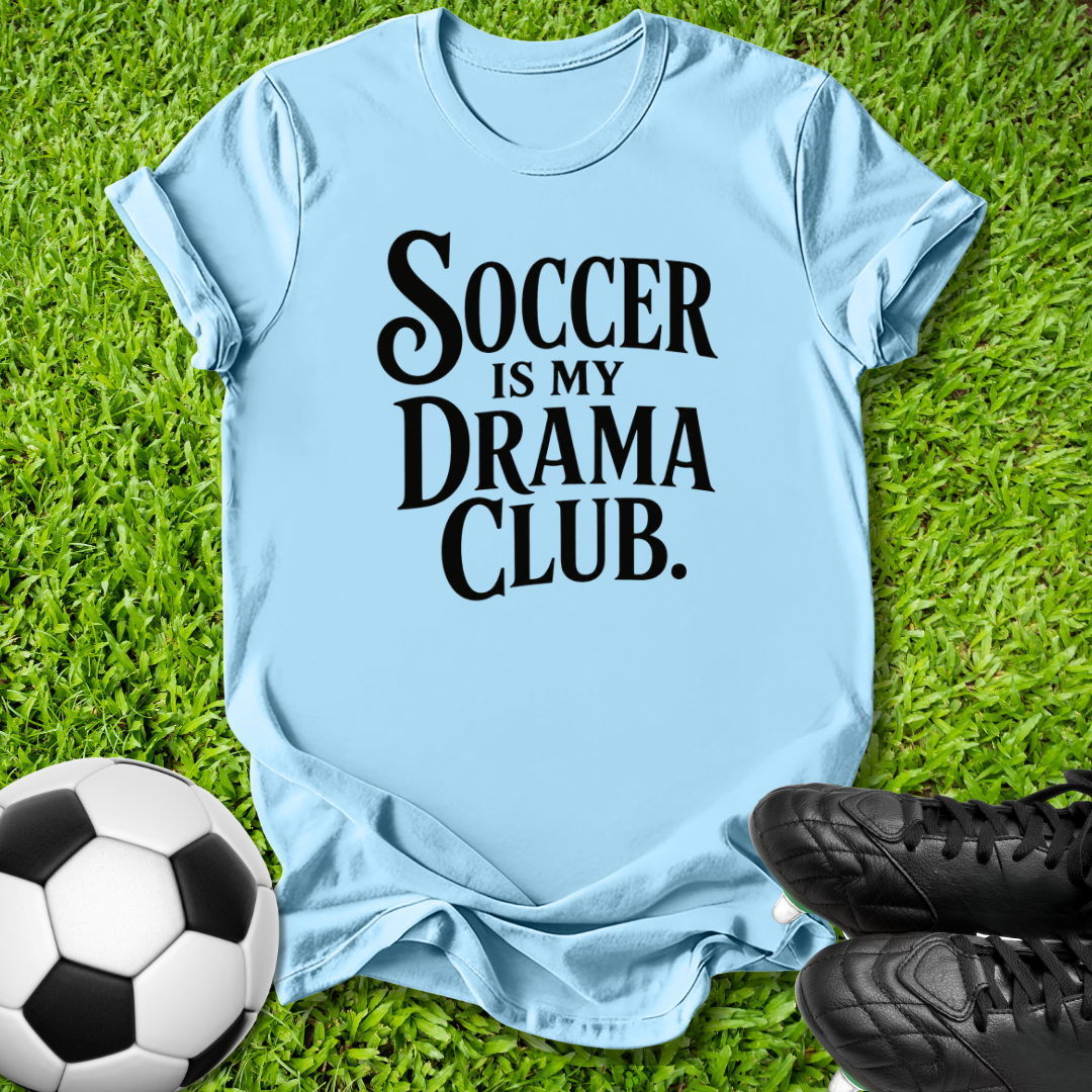 A light blue t-shirt with the text 'SOCCER IS MY DRAMA CLUB.' printed in black, laid flat on green grass with a soccer ball and black cleats as props.