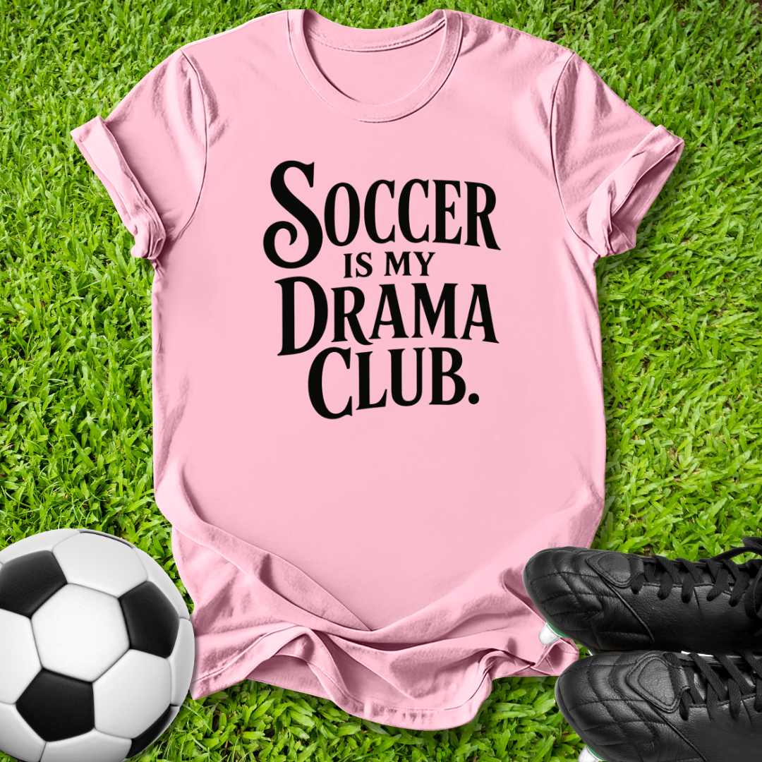 A pink short-sleeve t-shirt with the text 'SOCCER IS MY DRAMA CLUB.' printed in black, laid flat on green grass with a soccer ball and black cleats as props.