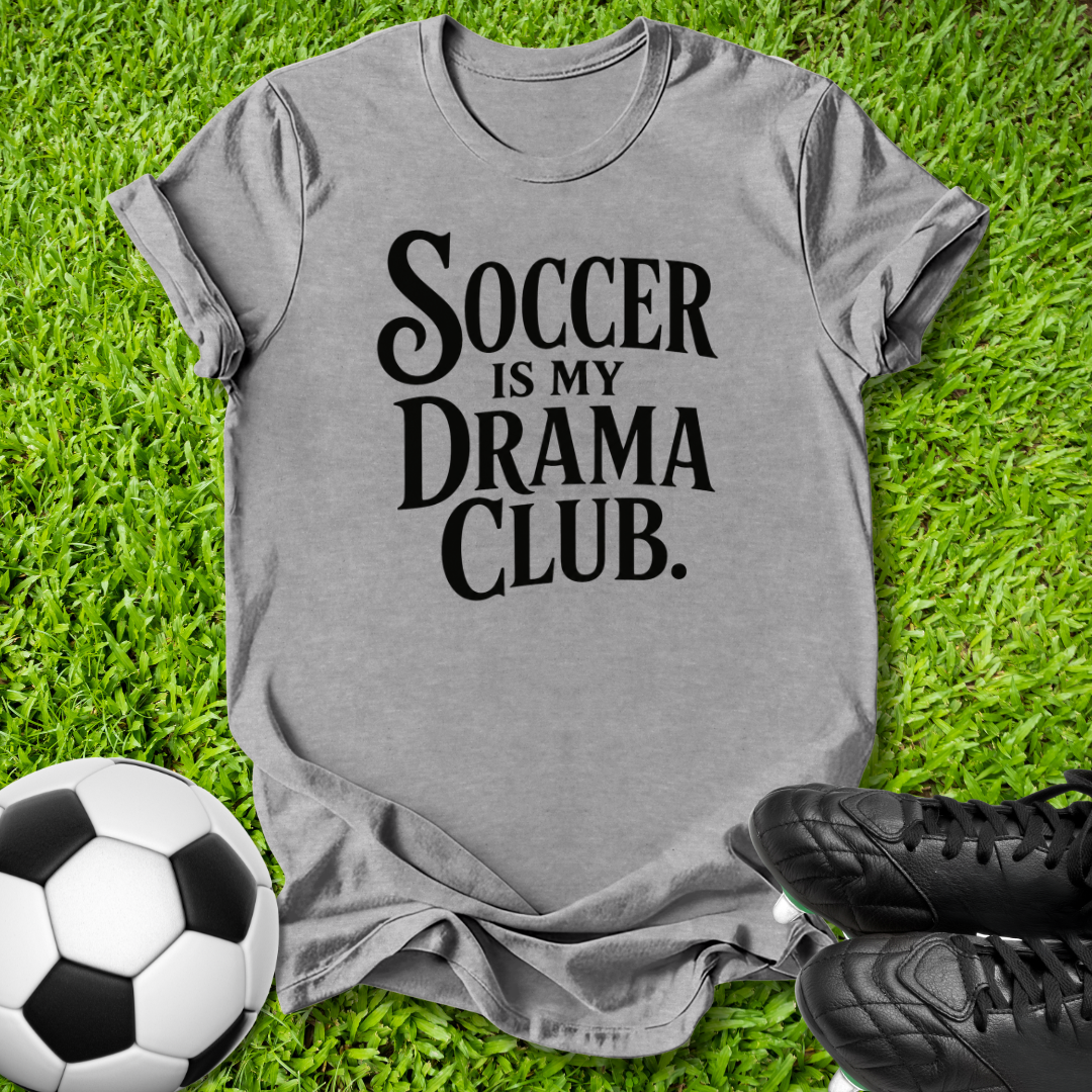 A heather gray short-sleeve t-shirt with the text 'SOCCER IS MY DRAMA CLUB.' printed in black across the front, laid flat on green grass with a soccer ball and black cleats nearby.