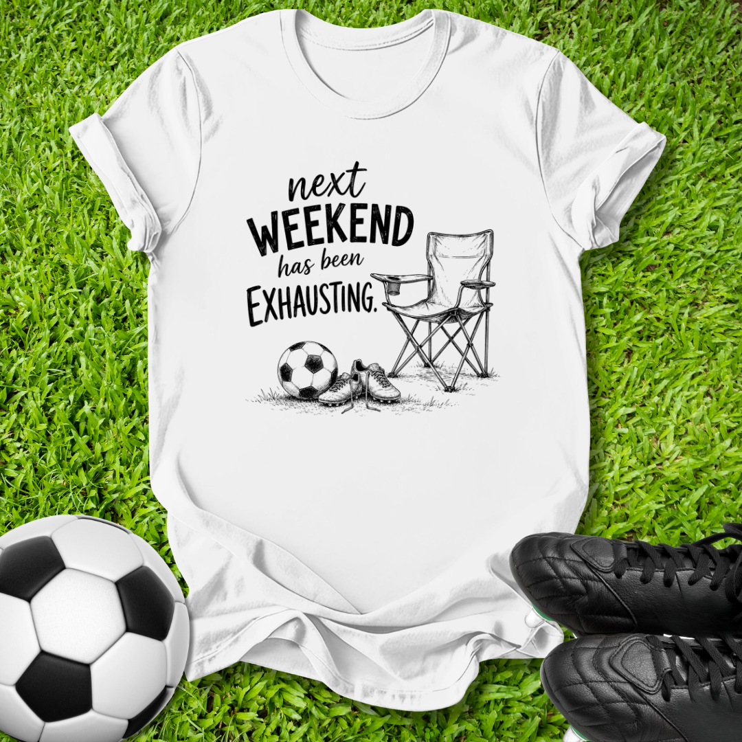 A white graphic t-shirt laid flat on green grass, featuring a black hand-drawn style print that reads 'next WEEKEND has been Exhausting' alongside a folding chair, soccer ball, and athletic shoes. A real soccer ball and black cleats are placed around the shirt as props.