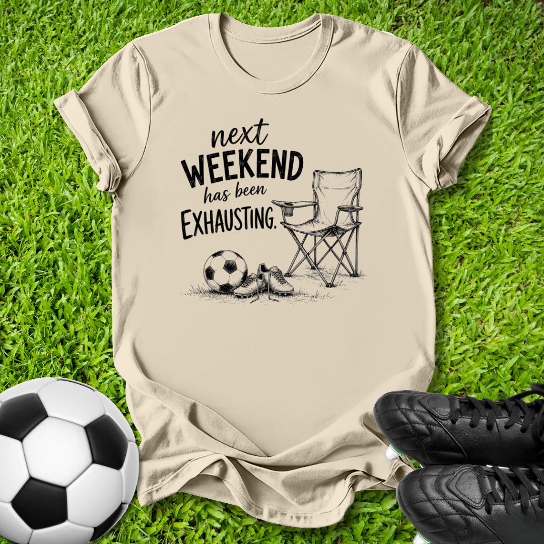 A beige graphic t-shirt laid flat on green grass, featuring the text 'next WEEKEND has been Exhausting' alongside a sketch of a folding chair, soccer ball, and cleats. A real soccer ball and black cleats are placed as props in the bottom corners.