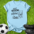 A light blue graphic t-shirt laid flat on green grass, featuring a black printed design of a folding chair, soccer ball, and shoes with the text 'next WEEKEND has been Exhausting.' Soccer balls and black cleats are placed around the shirt as props.