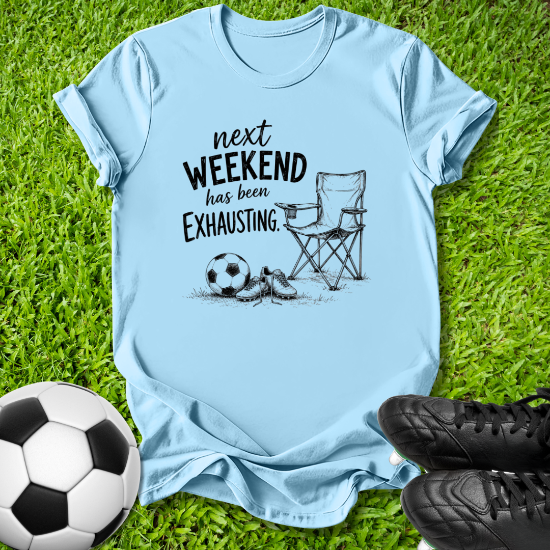 A light blue graphic t-shirt laid flat on green grass, featuring a black printed design of a folding chair, soccer ball, and shoes with the text 'next WEEKEND has been Exhausting.' Soccer balls and black cleats are placed around the shirt as props.