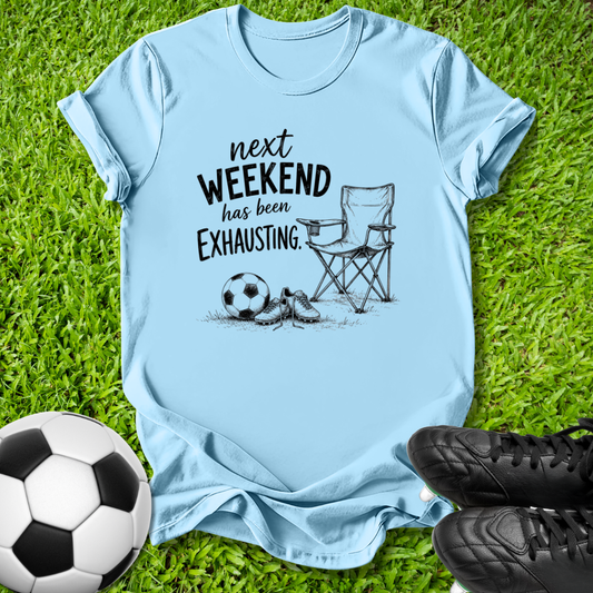 A light blue graphic t-shirt laid flat on green grass, featuring a black printed design of a folding chair, soccer ball, and shoes with the text 'next WEEKEND has been Exhausting.' Soccer balls and black cleats are placed around the shirt as props.