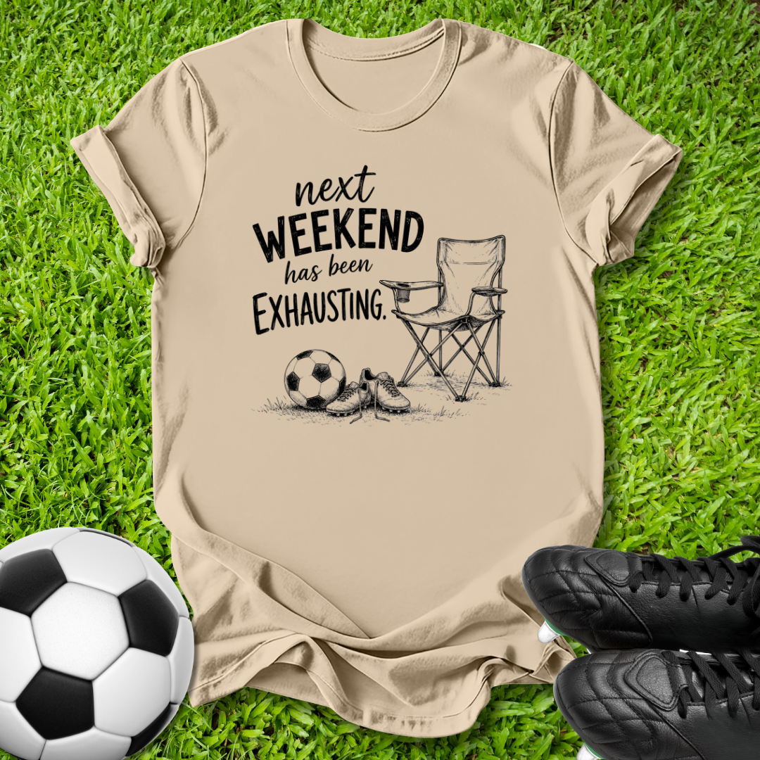 A beige graphic t-shirt laid flat on green grass, featuring a black print that reads 'next WEEKEND has been Exhausting' alongside a folding chair, soccer ball, and cleats. A soccer ball and black cleats are placed in the foreground as props.