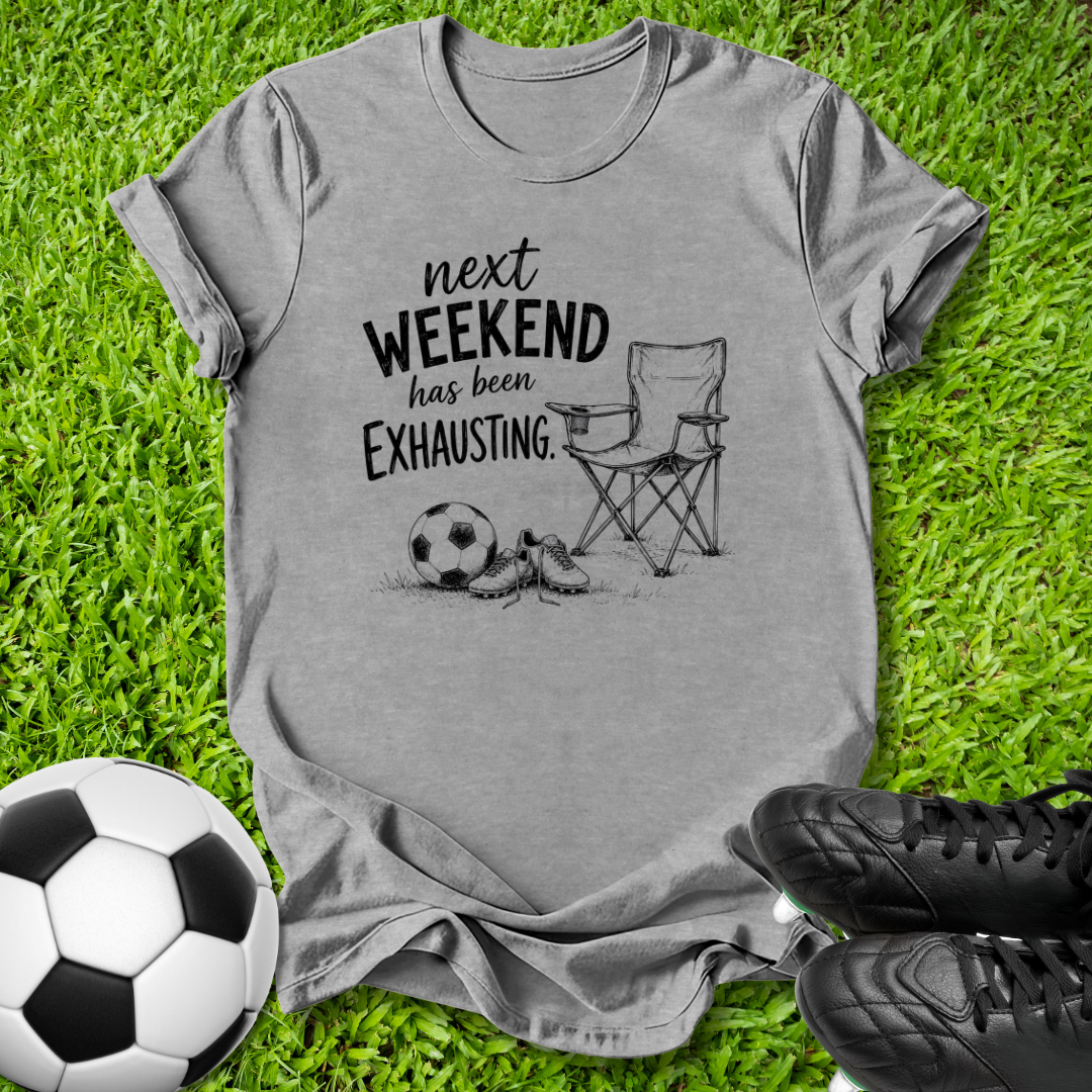 A heather gray graphic t-shirt laid flat on green grass, featuring a black printed design of a folding chair, a soccer ball, and a pair of cleats with the text 'next WEEKEND has been Exhausting.' Soccer shoes and a soccer ball are styled around the shirt.