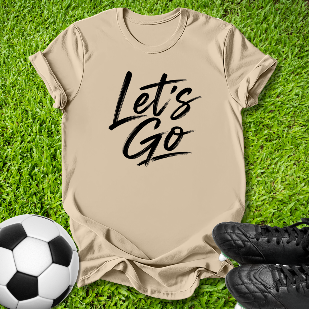 A beige short-sleeve T-shirt laid flat on green grass, with black script text reading 'Let's Go' across the chest. A soccer ball appears at the lower left, and black cleats are at the lower right, creating a sports-themed flat lay.