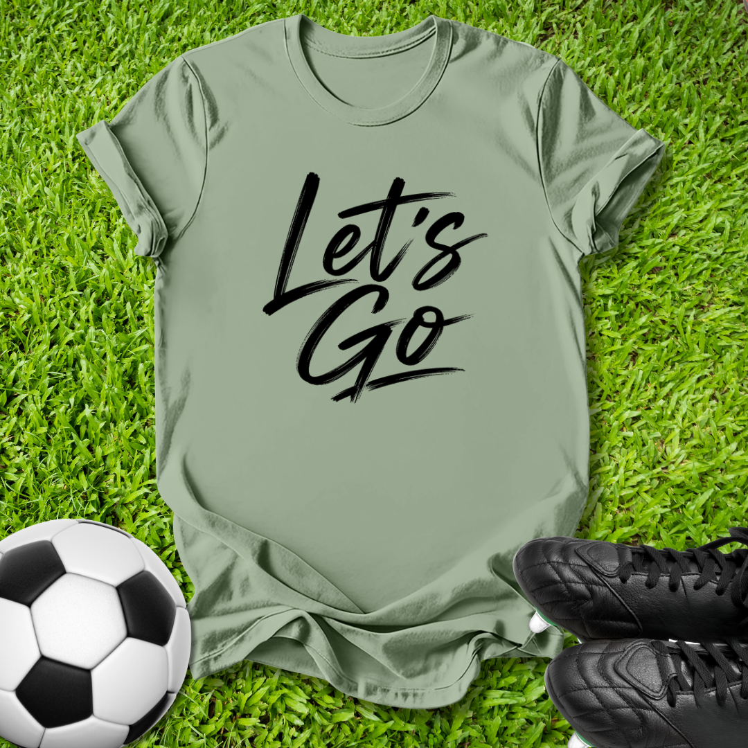 An olive green t-shirt with 'Let's Go' printed in black script across the chest, laid flat on green grass with a soccer ball and black cleats as props.