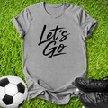 A heather gray short-sleeve T-shirt with the words 'Let's Go' printed in black script across the chest, laid flat on green grass with a soccer ball and black cleats nearby.