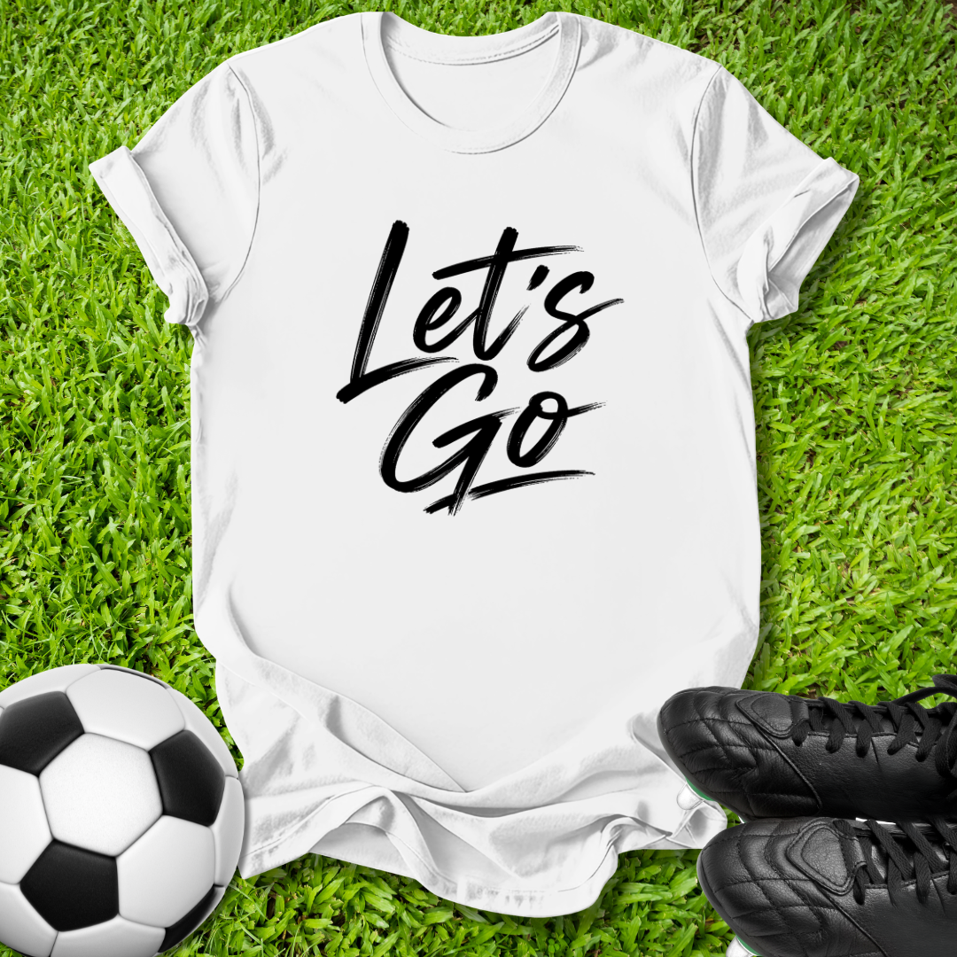 A white short-sleeve T-shirt laid flat on green grass, featuring the words 'Let's Go' in black script across the chest. A soccer ball appears at the lower left, and black cleats are at the lower right, creating a sports-themed flat lay.