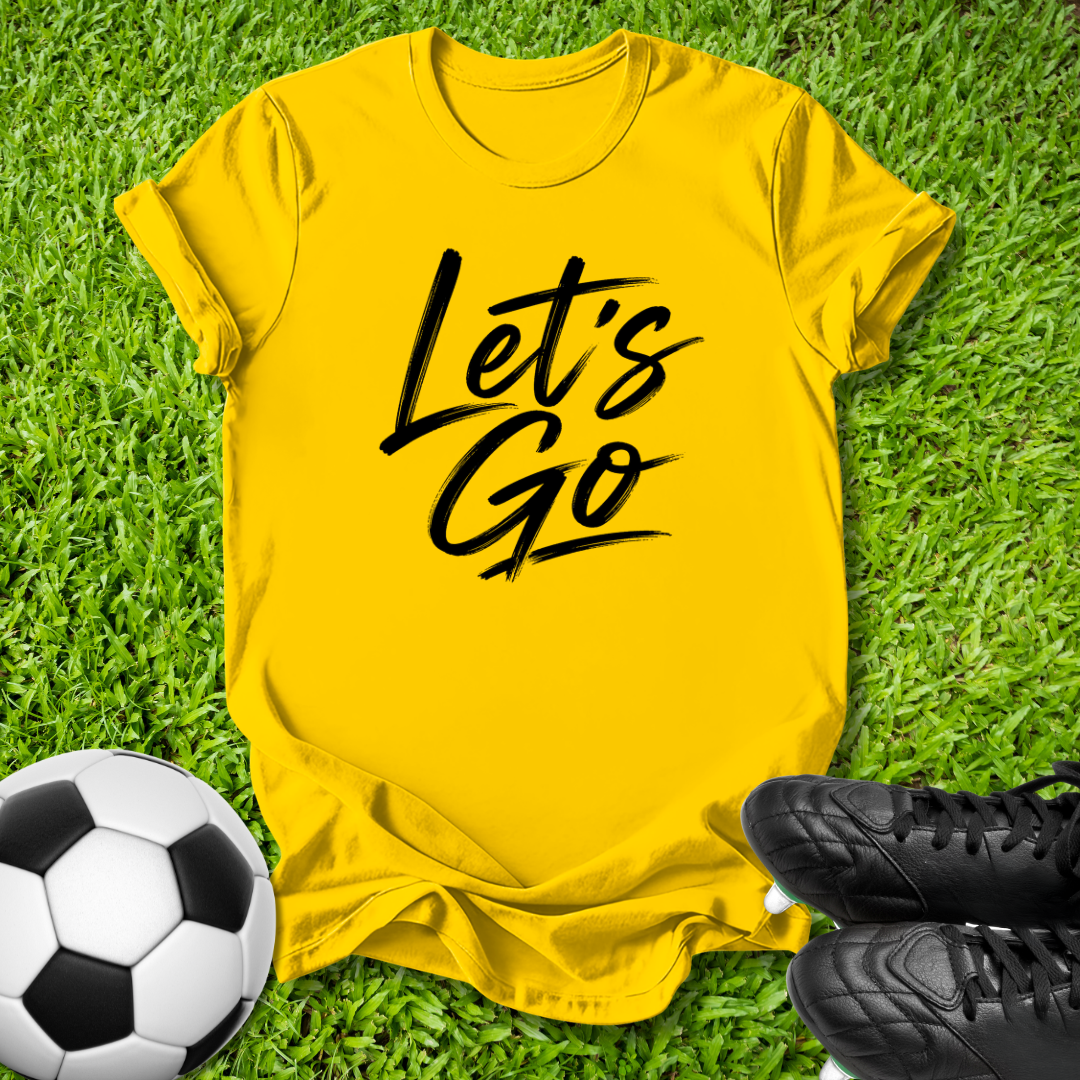 A bright yellow short-sleeve t-shirt with 'Let's Go' printed in black script across the chest, laid flat on green grass with a soccer ball and black cleats as props.
