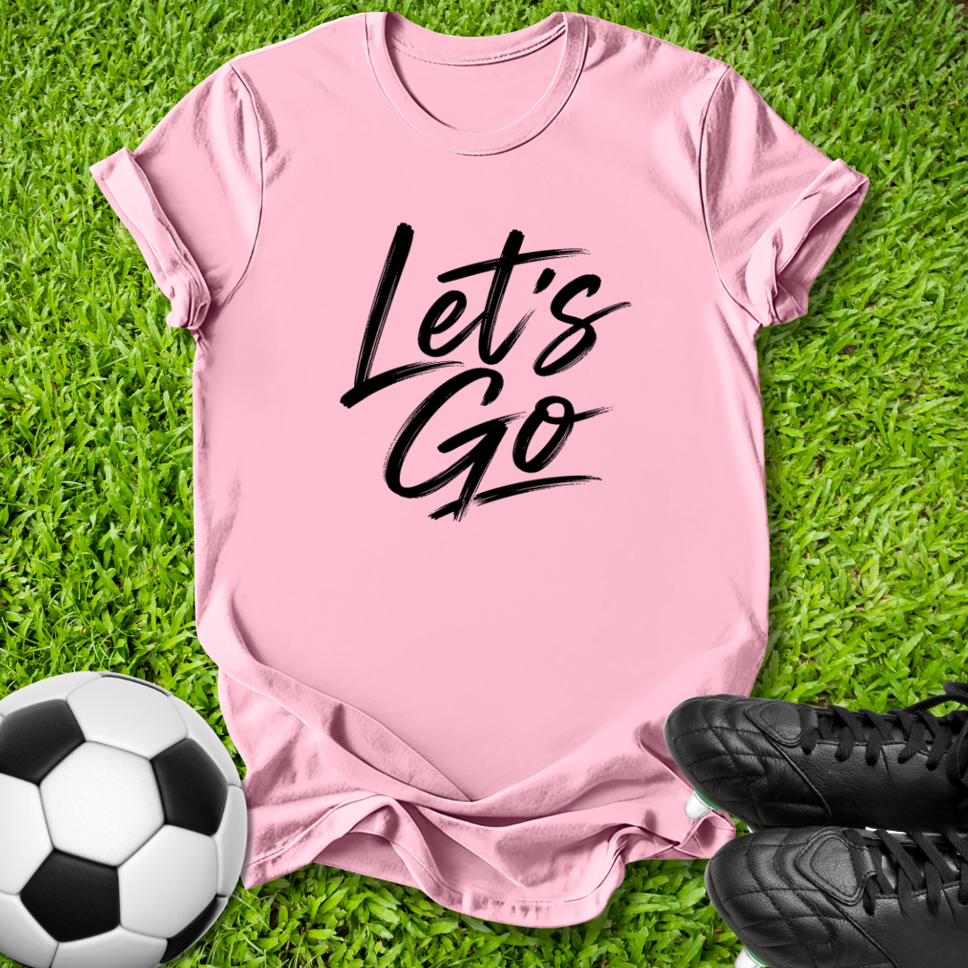 A pink short-sleeve t-shirt with 'Let's Go' printed in black script across the chest, laid flat on green grass with a soccer ball and black cleats as props.