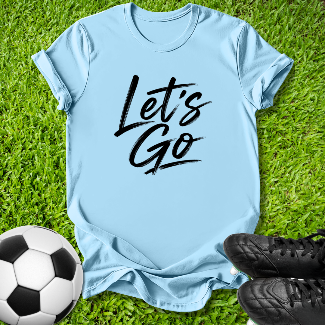A light blue short-sleeve t-shirt with 'Let's Go' printed in black script across the chest, laid flat on green grass with a soccer ball and black cleats as props.