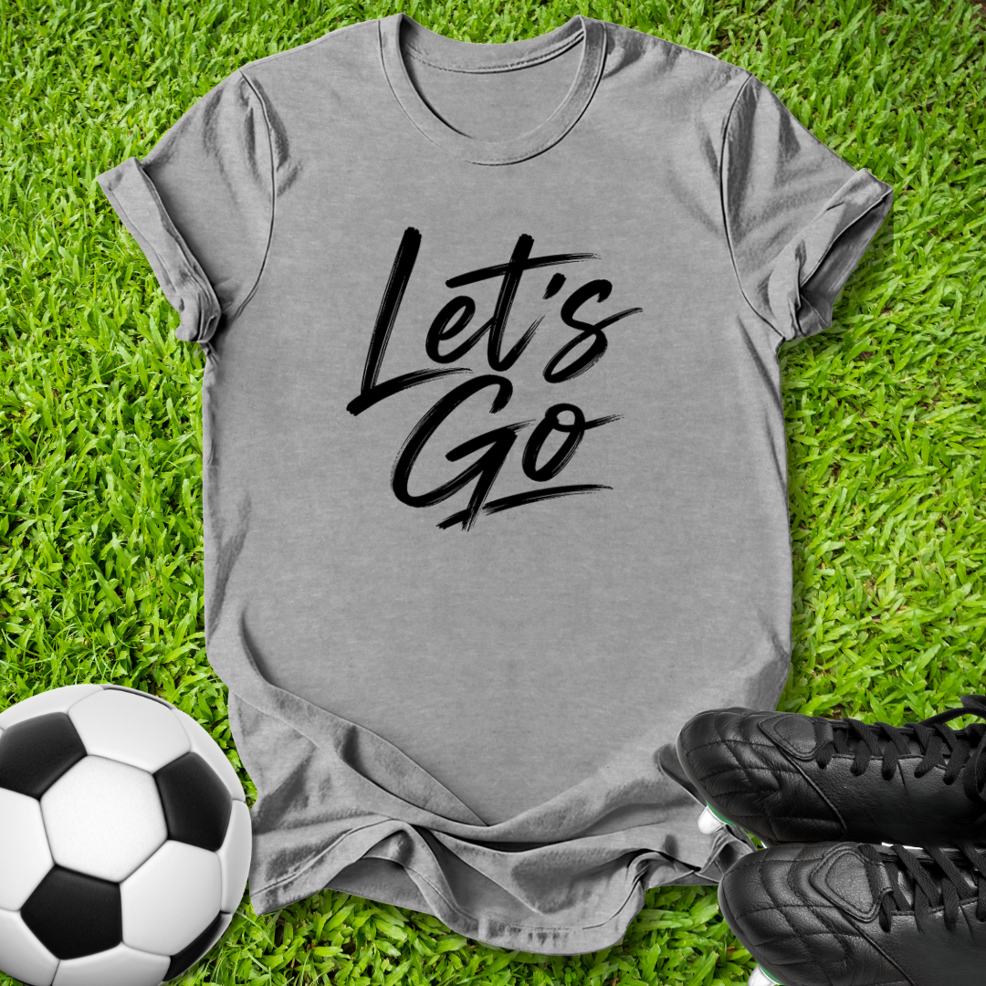 A heather gray short-sleeve T-shirt with the words 'Let's Go' printed in black script across the chest, laid flat on green grass with a soccer ball and black cleats nearby.
