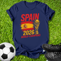 A navy blue t-shirt laid flat on green grass, featuring a distressed red, yellow, and gold graphic that reads 'SPAIN 2026 WORLD CHAMPIONS' with the FIFA trophy illustration. A soccer ball is visible on the lower left and black cleats on the lower right.