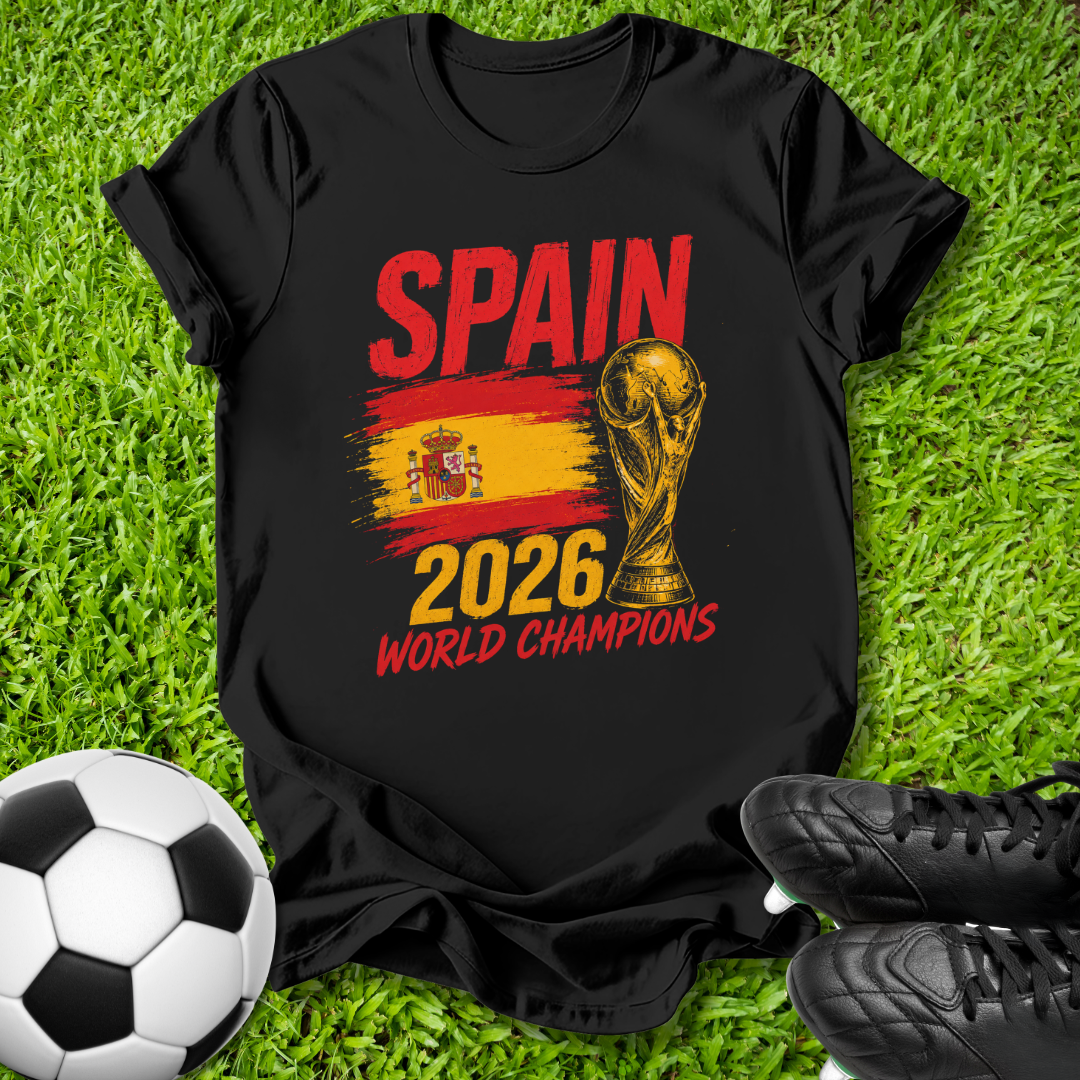 A black short-sleeve graphic T-shirt laid flat on green grass, featuring the word 'SPAIN' in red, a Spanish flag stripe, the FIFA World Cup trophy, and '2026 WORLD CHAMPIONS' in yellow and red. A soccer ball and black cleats are placed around it as styling props.