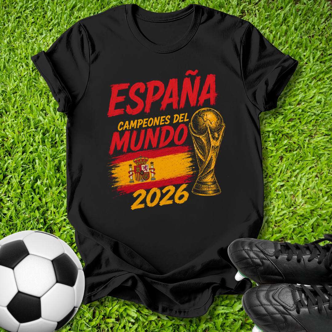 A black graphic t-shirt laid flat on green grass, featuring red and yellow text that reads 'ESPAÑA CAMPEONES DEL MUNDO 2026' with a trophy graphic and Spanish flag colors. A soccer ball and black cleats are placed around it as props.