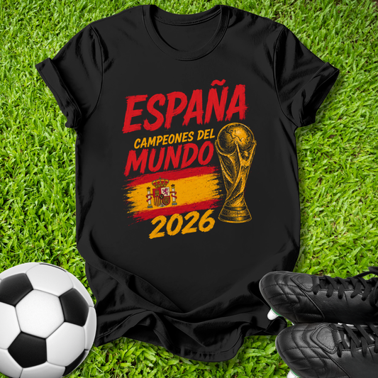 A black graphic t-shirt laid flat on green grass, featuring red and yellow text that reads 'ESPAÑA CAMPEONES DEL MUNDO 2026' with a trophy graphic and Spanish flag colors. A soccer ball and black cleats are placed around it as props.