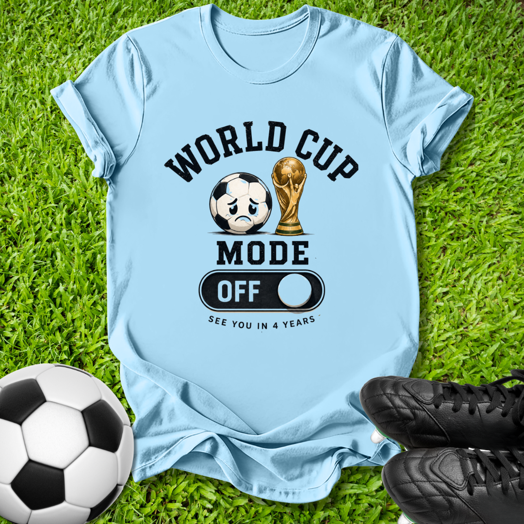 A light blue graphic t-shirt laid flat on green grass, featuring a soccer ball and trophy graphic with the text 'WORLD CUP MODE OFF' and 'SEE YOU IN 4 YEARS'. A black-and-white soccer ball, black cleats, and the grass background complete the scene.