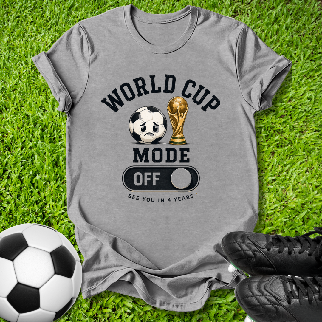 A heather gray graphic t-shirt laid flat on green grass, featuring a soccer-themed design with a soccer ball, trophy, and the words 'WORLD CUP MODE OFF' and 'SEE YOU IN 4 YEARS'. A soccer ball and black cleats are placed around it as props.