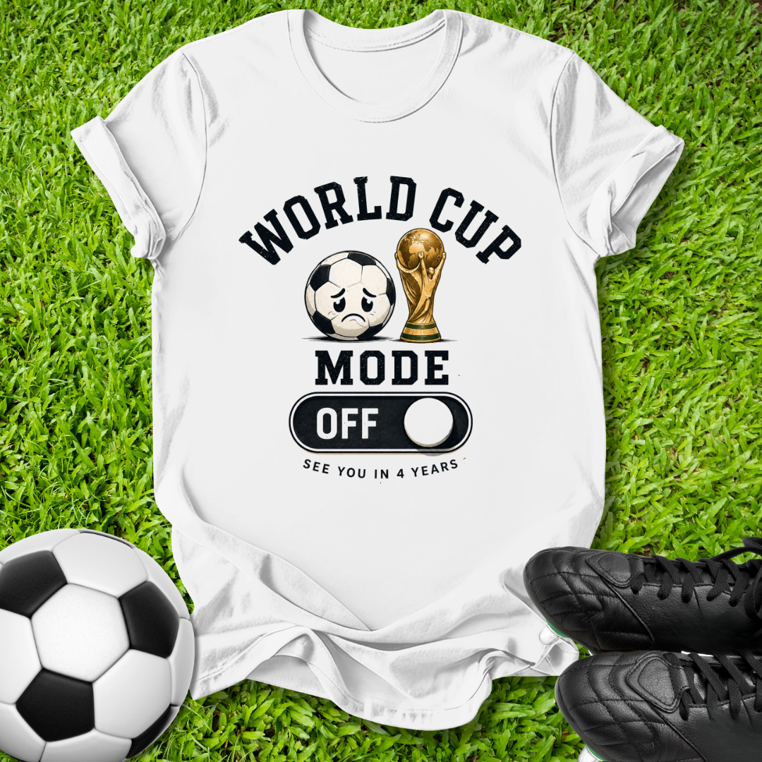 A white graphic t-shirt laid flat on green grass, featuring a black and gold World Cup-themed design with a soccer ball, trophy, and the text 'WORLD CUP MODE OFF SEE YOU IN 4 YEARS'. A black-and-white soccer ball is at the bottom left, and black cleats are at the bottom right.