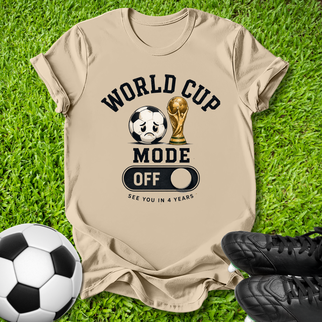 A beige graphic t-shirt laid flat on green grass, featuring a World Cup-themed design with a soccer ball, trophy, and the text 'WORLD CUP MODE OFF SEE YOU IN 4 YEARS'. A soccer ball and black cleats are placed around it as props.