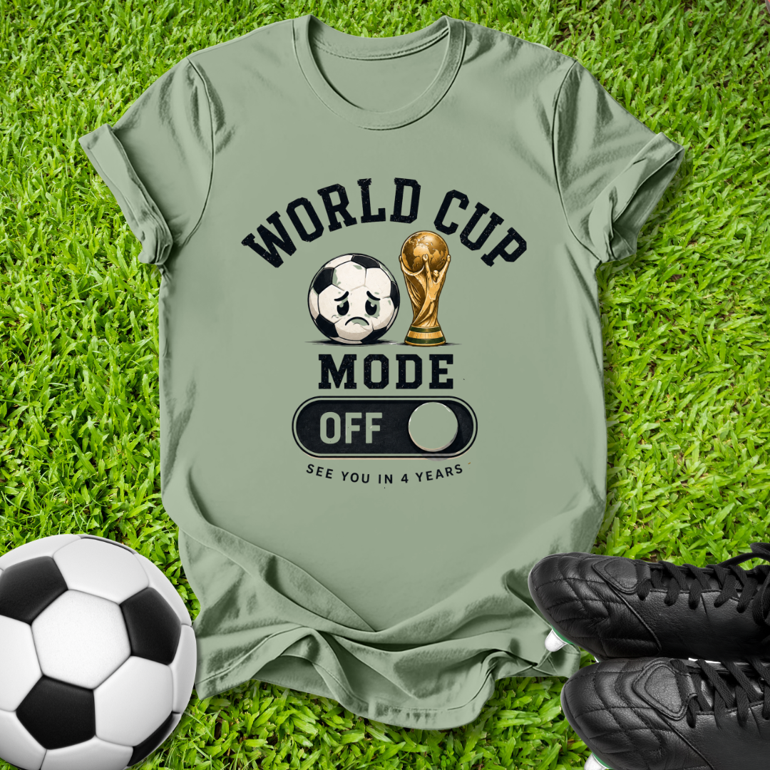 An olive green graphic t-shirt laid flat on green grass, featuring a 'WORLD CUP MODE OFF' design with a soccer ball and trophy graphic. A black-and-white soccer ball and black cleats are placed around it as props.