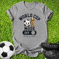 A heather gray graphic t-shirt laid flat on green grass, featuring a soccer-themed design with a soccer ball, trophy, and the words 'WORLD CUP MODE OFF' and 'SEE YOU IN 4 YEARS'. A soccer ball and black cleats are placed around it as props.
