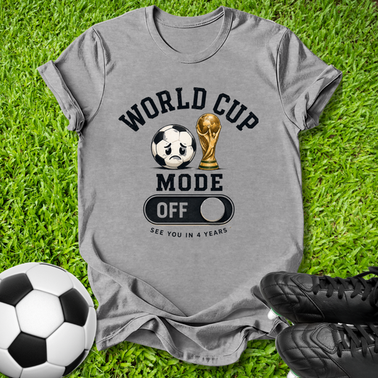 A heather gray graphic t-shirt laid flat on green grass, featuring a soccer-themed design with a soccer ball, trophy, and the words 'WORLD CUP MODE OFF' and 'SEE YOU IN 4 YEARS'. A soccer ball and black cleats are placed around it as props.